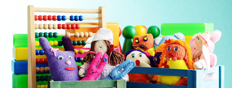 Eine Nahaufnahme verschiedener Spielzeuge in kleinen bunten Holzkisten. Man sieht Stoffpuppen, eine Gummiente, Spielfiguren und einen Abakus mit bunten Holzperlen.