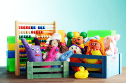 Eine Nahaufnahme verschiedener Spielzeuge in kleinen bunten Holzkisten. Man sieht Stoffpuppen, eine Gummiente, Spielfiguren und einen Abakus mit bunten Holzperlen.