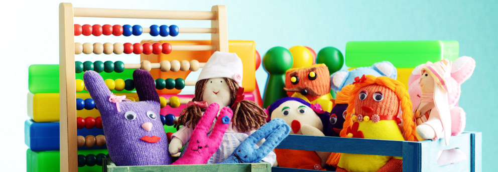 Eine Nahaufnahme verschiedener Spielzeuge in kleinen bunten Holzkisten. Man sieht Stoffpuppen, eine Gummiente, Spielfiguren und einen Abakus mit bunten Holzperlen.
